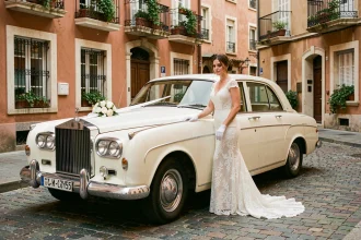 Mariée en robe de dentelle posant à côté d'une Rolls-Royce vintage crème décorée d'un ruban blanc dans une rue pavée de Toulouse